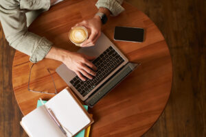co working space, freelance and e learning concept. top view, male hands holding cup of coffee and using laptop. a man sitting in cafe and working or studying online