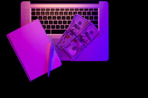 laptop, blank notepad and paper money in neon lighting, top view.